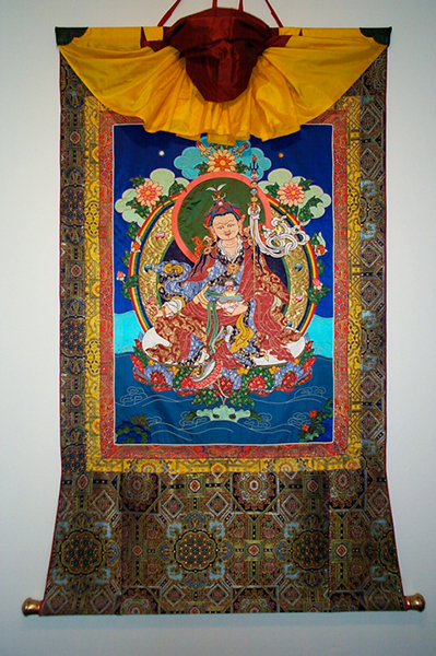 Guru Rinpoche appliqué thangka by Leslie Rinchen-Wongmo. Notice the "rainbow" strips of brocade surrounding the image, the slat at the top of the frame, dowel at the bottom, and veil decoratively arranged at the top.